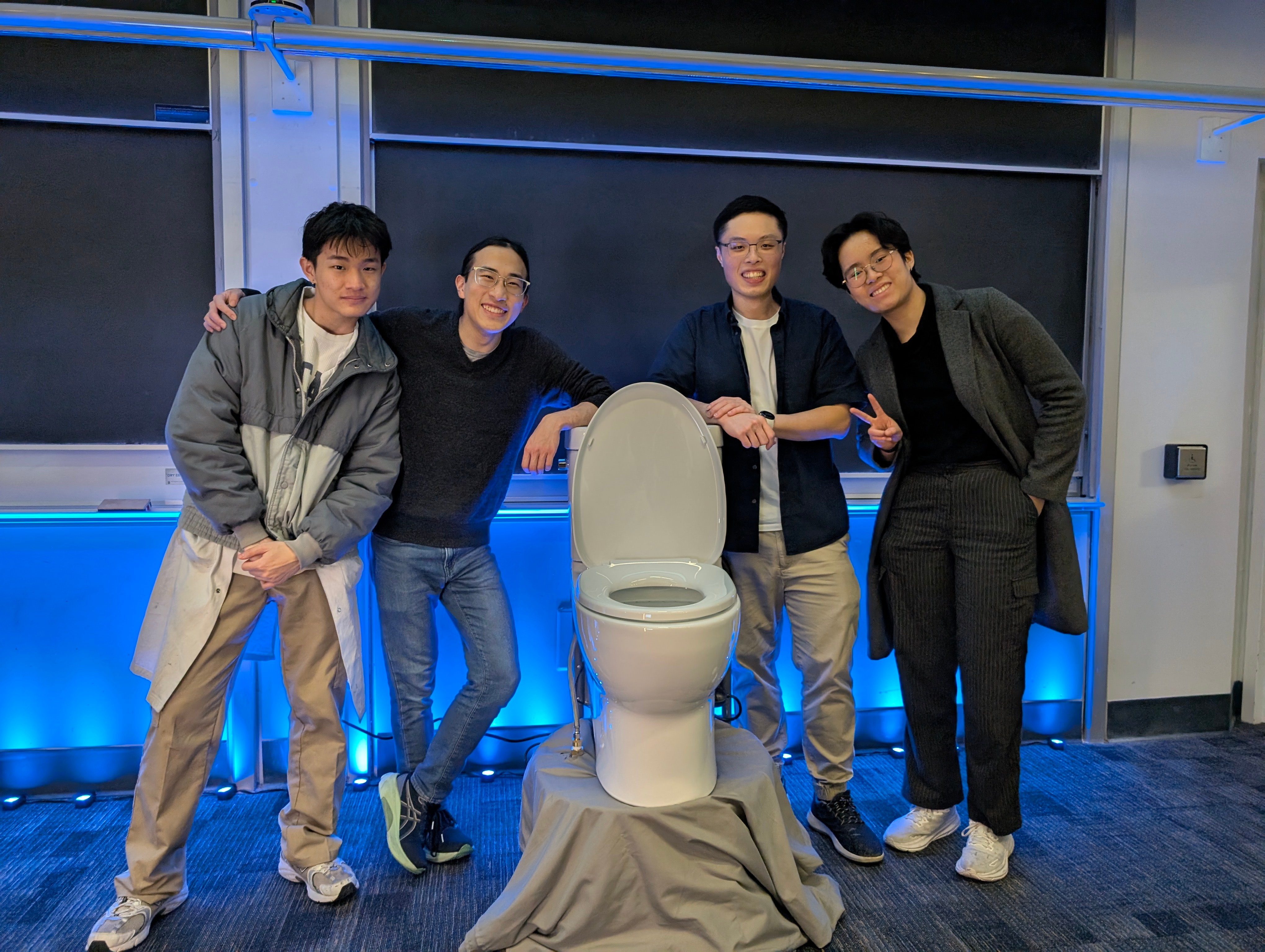 Four young adults—Bill, Kai, Calvin, and Chloe—pose indoors in front of a toilet mounted on a small platform draped with gray fabric. They are smiling and casually dressed; Chloe, on the far right, is flashing a peace sign. The background features a classroom setting with large blackboards and blue accent lighting along the wall. The atmosphere is playful and lighthearted.